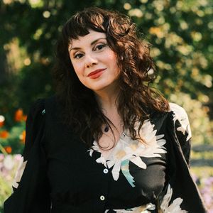 Portrait of author Gina María Balibrera standing outdoors in a lush, sun-dappled garden. She has wavy dark brown hair with short bangs and wears a black dress with large white floral prints. Her expression is calm and confident, with a gentle smile and warm red lipstick. Soft sunlight highlights her face and hair, and a blurred background of greenery and blooming flowers adds a natural, serene ambiance.